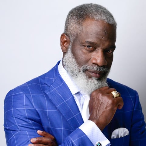 Older man with gray beard wearing a blue plaid blazer and white dress shirt, posed thoughtfully against a light background